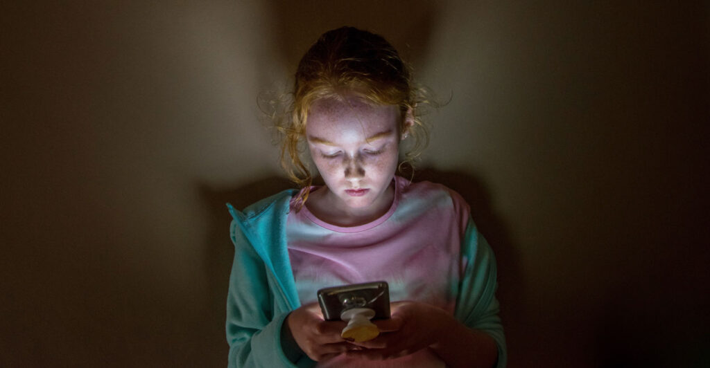Pale young girl, her face lit by the light of her cellphone, looks down at the screen.