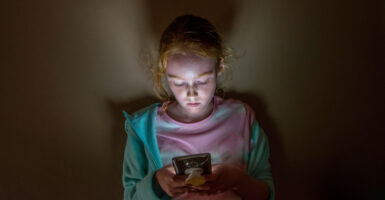 Pale young girl, her face lit by the light of her cellphone, looks down at the screen.