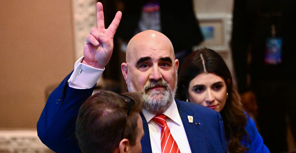 Trump 2024 campaign co-chair Chris LaCivita in blue suit and red tie flashes peace sign.