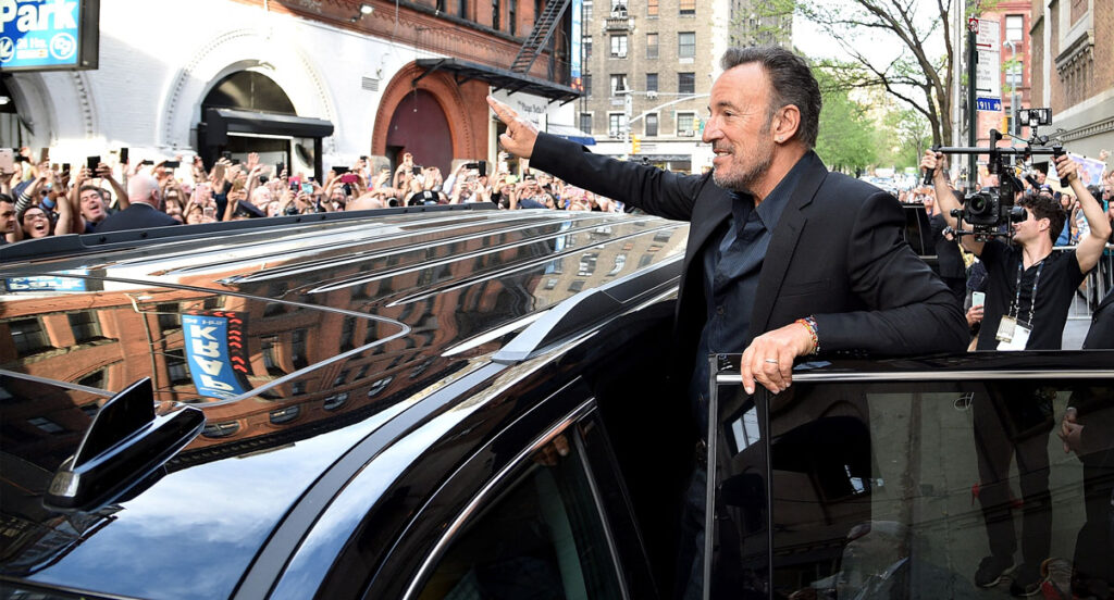 Standing atop the doorway of a black limousine, Bruce Springsteen waves to a crowd of fans standing across the street.