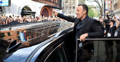 Standing atop the doorway of a black limousine, Bruce Springsteen waves to a crowd of fans standing across the street.