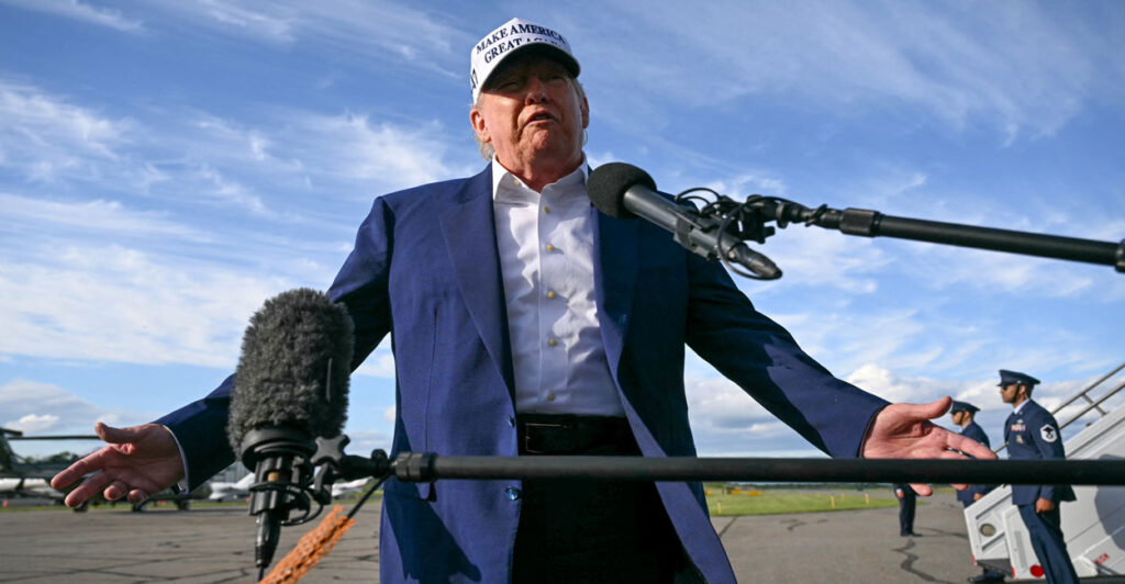 President Donald Trump, in a white "Make America Great Hat" addresses reporters, with his arms outstretched, on the tarmac of a New Jersey airport.