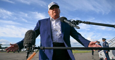 President Donald Trump, in a white "Make America Great Hat" addresses reporters, with his arms outstretched, on the tarmac of a New Jersey airport.