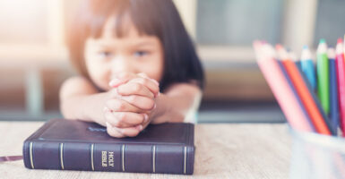 Brown-haired little girl with her hands clasped on top of a Bible. A bunch of crayons to her left.