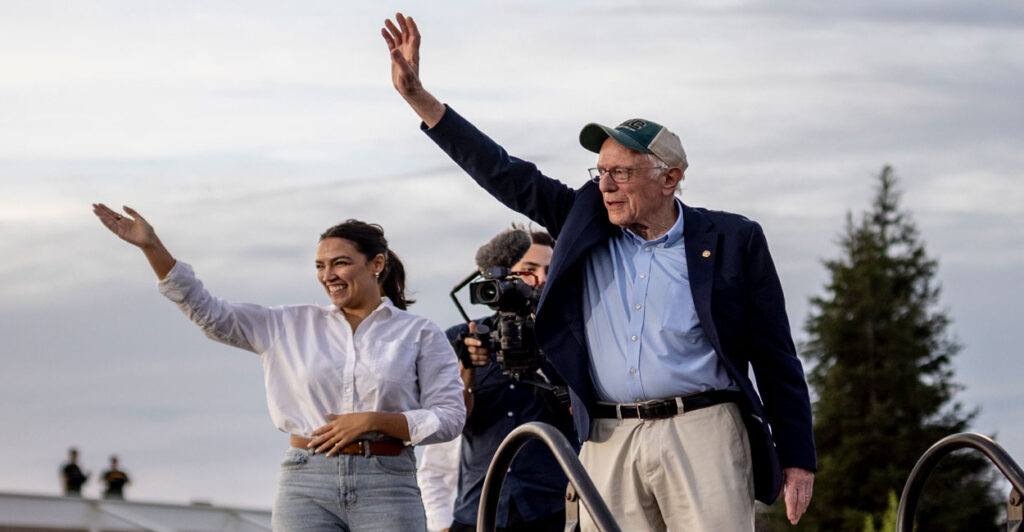 Bernie Sanders in blue jacket and hat, along with Alexandria Ocasio-Cortez in white shirt and jeans wave to crowd gathered for a "Fighting Oligarchy" rally.
