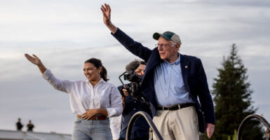 Bernie Sanders in blue jacket and hat, along with Alexandria Ocasio-Cortez in white shirt and jeans wave to crowd gathered for a "Fighting Oligarchy" rally.