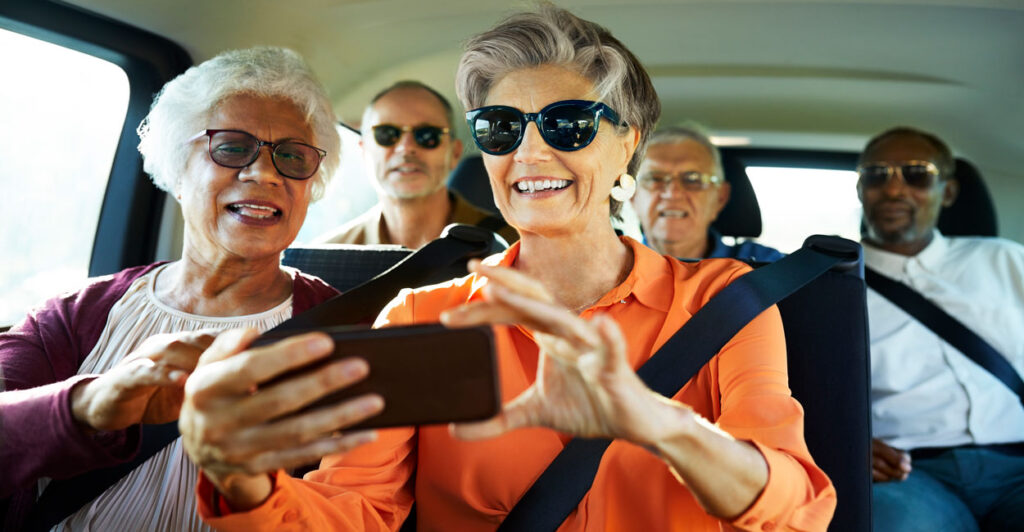 A group of senior citizens of mixed races happy ride in a minivan. The two ladies in the front look at a smartphone.