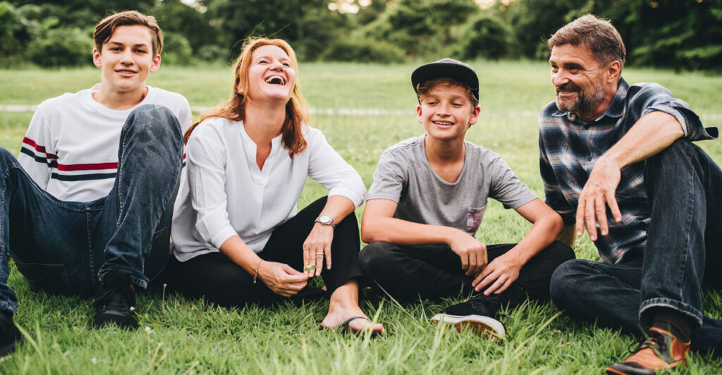 A mother and father with their two sons sit on grass, with the mother laughing.