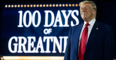 Donald Trump in a blue suit and red tie standing next to a backdrop banner that says 100 days of greatness