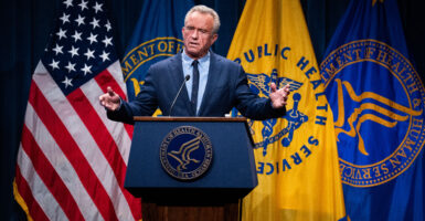 Behind a lectern, Health and Human Services Secretary Robert F. Kennedy Jr. gestures during remarks, with U.S., HHS, and Public Health Service flags displayed behind him.