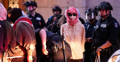 A masked protester in colorful blouse in police custody. Behind the suspect a row of heavily armored police. In front, an officer bends down.