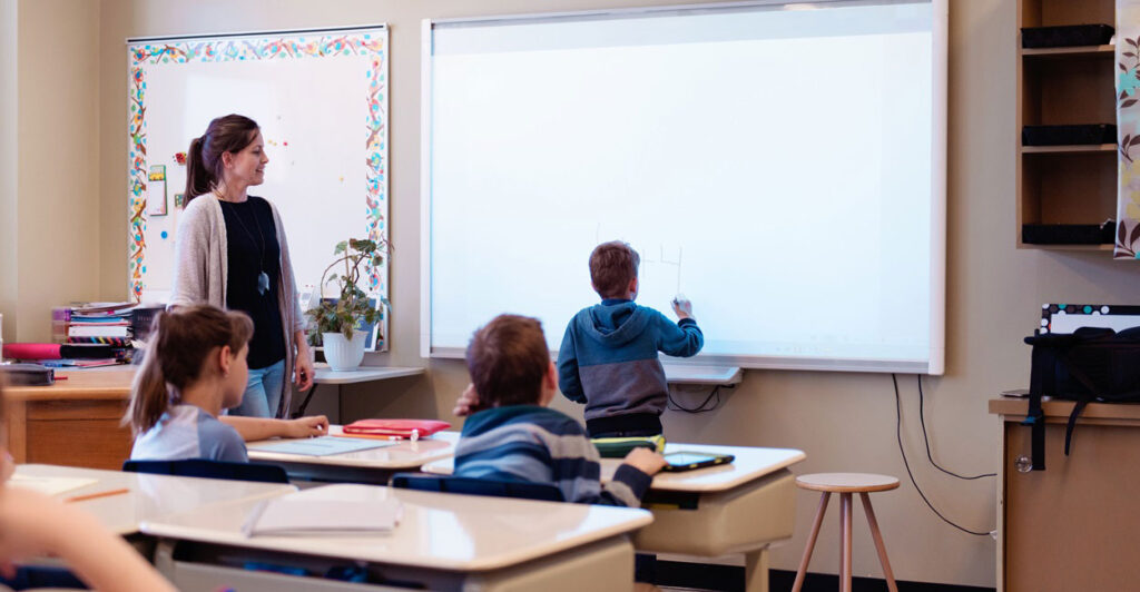 A teacher stands to the left of a child and observes while the child writes on the chalkboard.