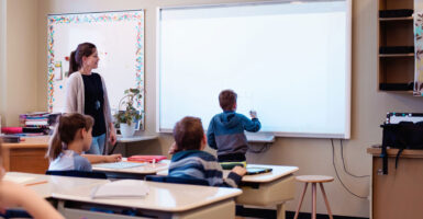 A teacher stands to the left of a child and observes while the child writes on the chalkboard.