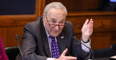 Chuck Schumer sits at a table with a microphone in front of him and talks while gesturing with his hands.