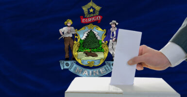 A hand deposits a ballot in a ballot box, with the Maine state flag in the background.