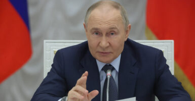 Vladimir Putin sits at a table behind a microphone and gestures with his right hand as he talks.