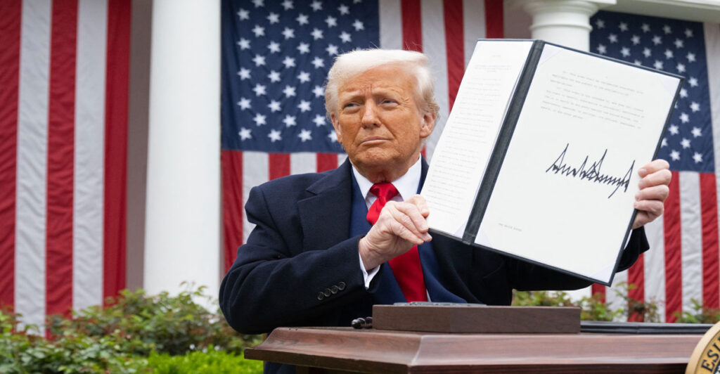 President Donald Trump in front of a large American flag on the White House lawn, sits at a desk, holding up a signed executive order.