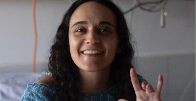Emily Damari smiles in a hospital bed and holds up her hand showing that she lost her middle and ring finger on her left hand.