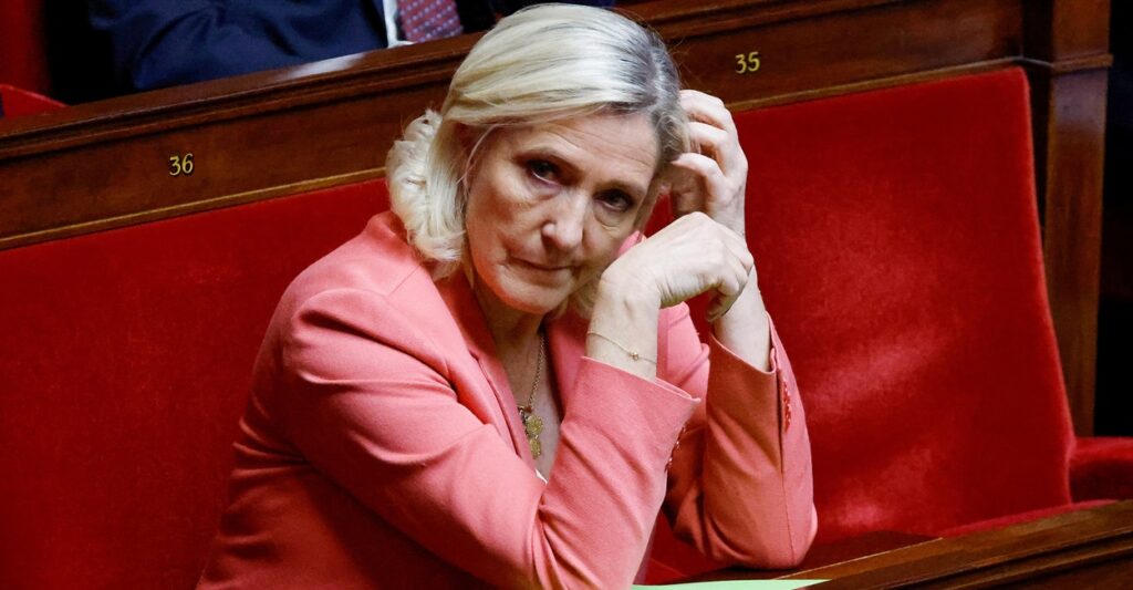 Marine Le Pen sits at a table with her hands near her face as her left hand touches her head.