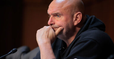 Senator John Fetterman sitting at a hearing in a hoodie with his chin resting on his hand