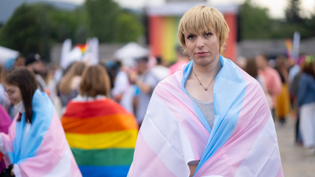 A gender-diverse person wearing a pansexual flag, standing in front of a crowd at a gay pride event.