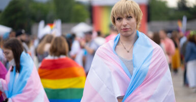 A gender-diverse person wearing a pansexual flag, standing in front of a crowd at a gay pride event.