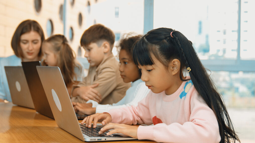 Four young children of mixed races and sexes work at laptops as teacher sits at end of table looking on.