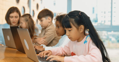 Four young children of mixed races and sexes work at laptops as teacher sits at end of table looking on.