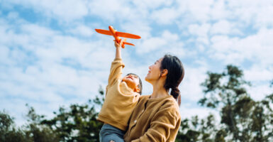 Young Asian mother and lovely little daughter spending time together outdoors, playing with airplane toy and smiling joyfully in park on a lovely sunny day against beautiful blue sky.