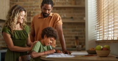 A black father and white, blonde mother affectionately stand over their son as he does homework.
