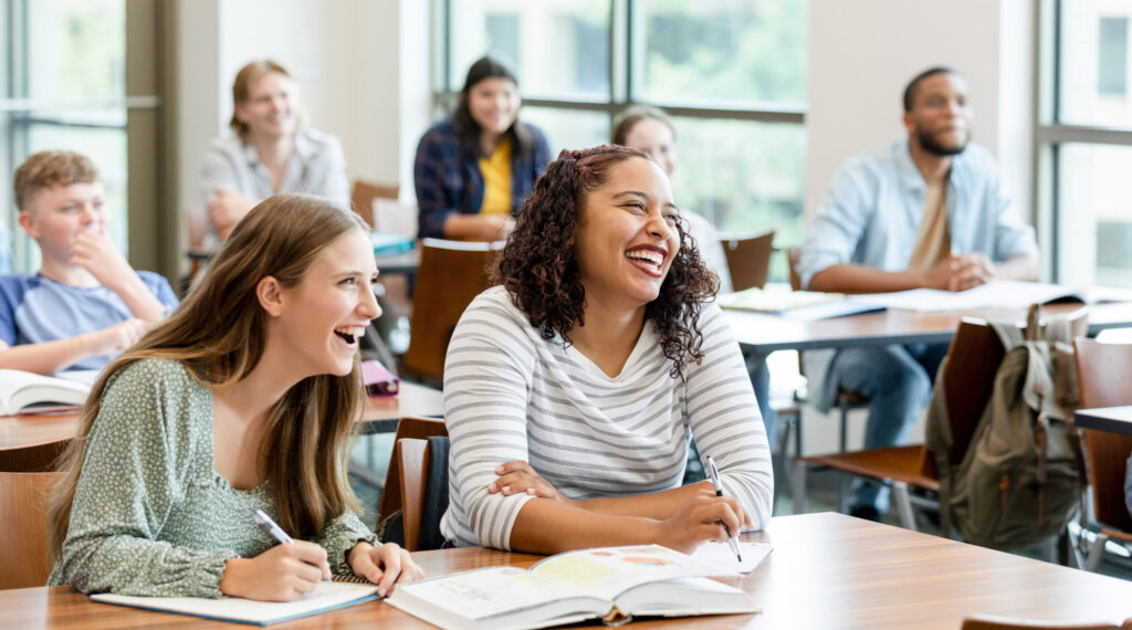 Two female college classmates at their desk laugh during a class. Four other students sit in background.