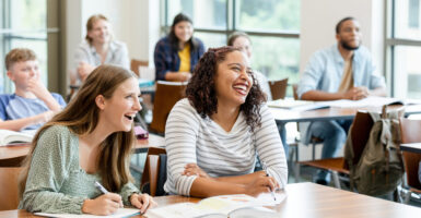 Two female college classmates at their desk laugh during a class. Four other students sit in background.