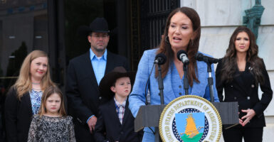Agriculture Secretary Brooke Rollins in a blue outfit speaks at a podium, with DHS Secretary Kristi Noem and the Maude family behind her.