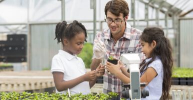 Inside a greenhouse, a white male botanist in glasses enthusiastically instructs a young black girl and young Hispanic girl. The Hispanic girl holds a small plant in a container that the botanist is probing.