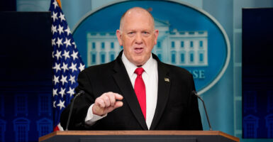 Tom Homan speaks from the podium of the White House press briefing room in a black suit and red tie.