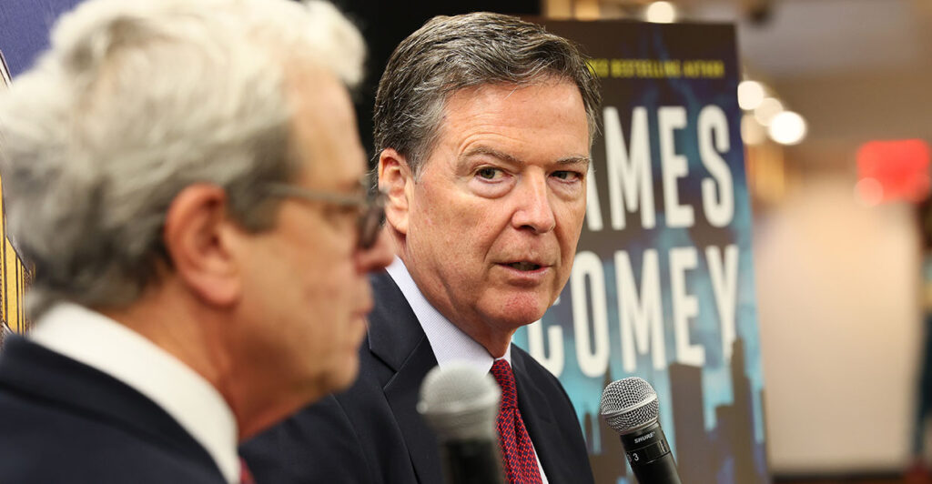 James Comey with a microphone in a suit in front of a book cover