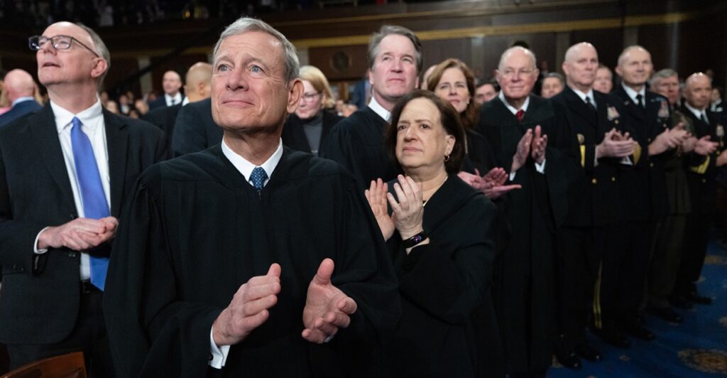 John Roberts is wearing his Supreme Court robe as he claps and looks to his right.