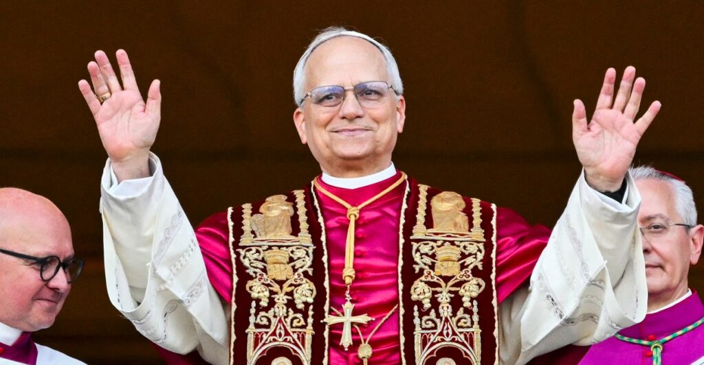 Pope Leo XIV stands with a smile on his face and holds his hands up in the air.