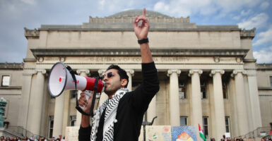 Mohsen Mahdawi in Palestinian garb with a megaphone and his hand raised in the air at a protest outside a Gothic structure stone building pillars on a university campus