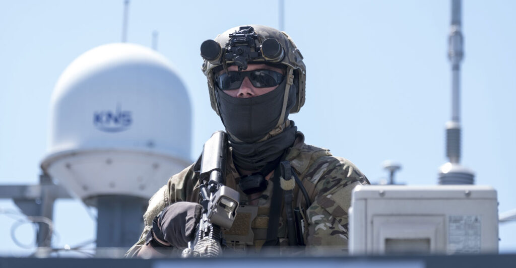 a Romanian soldier in military fatigues, a helmet, sunglasses, and a facemask holding an automatic weapon