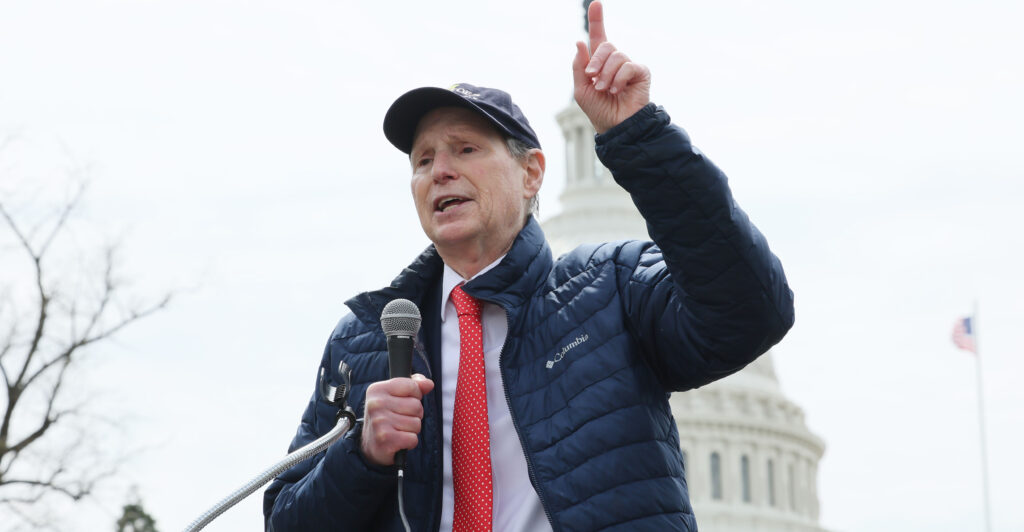 Sen. Ron Wyden, D-Ore., with the U.S. Capitol in the background, points upward while delivering a speech.