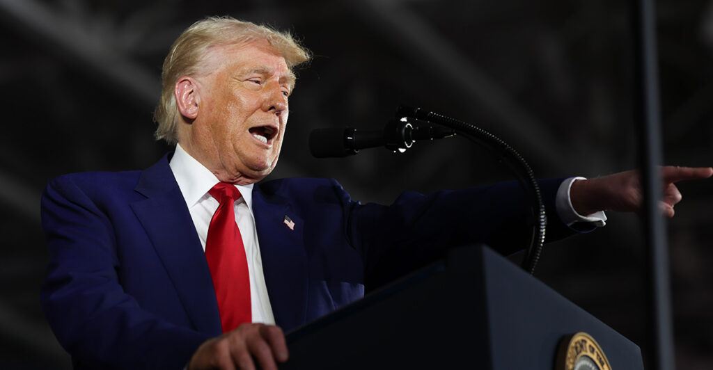 President Donald Trump in a blue suit and red tie speaks on stage at a podium