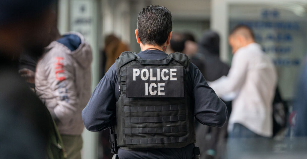 in immigration and customs enforcement agent stands in a room with his back to the camera in a bulletproof vest with a badge that says police ice on it
