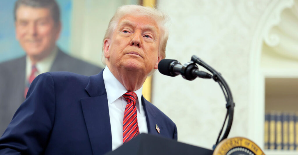 Donald Trump standing at a podium in the White House with a portrait of Ronald Reagan in the background