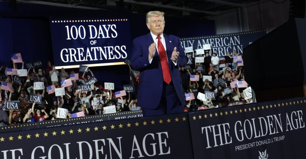 Donald Trump stands behind a banner and applauds while looking out at the crowd.