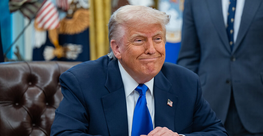 President Trump, hands folded sits at a desk, smiling a toothless smile.