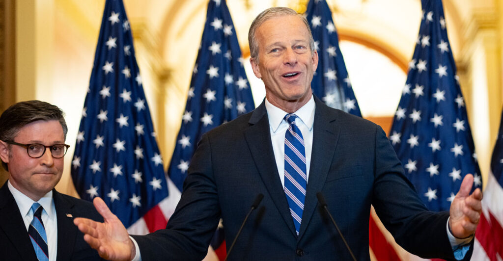 Mike Johnson stands next to John Thune, who has his arms spread wide. Behind the congressional leaders are four American flags.