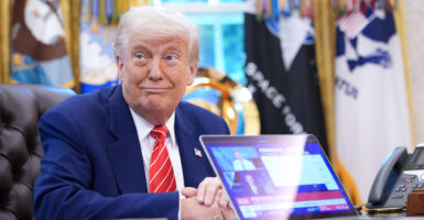 Donald Trump at desk in Oval Office, grinning. In front of him is a laptop, with screen facing camera.
