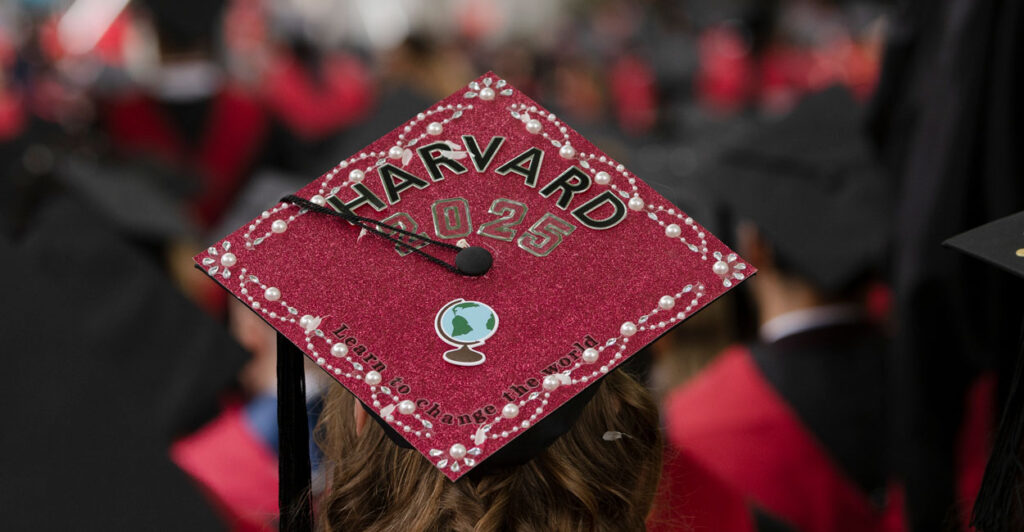A crimson Harvard graduation cap atop the head of a graduate. The hat reads "Harvard 2025" with a tassel crossing to the left, and in small letters "Learn to Change the World."