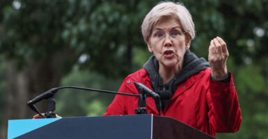 Sen. Elizabeth Warren in a red rain jacket speaks and gestures with her left hand.